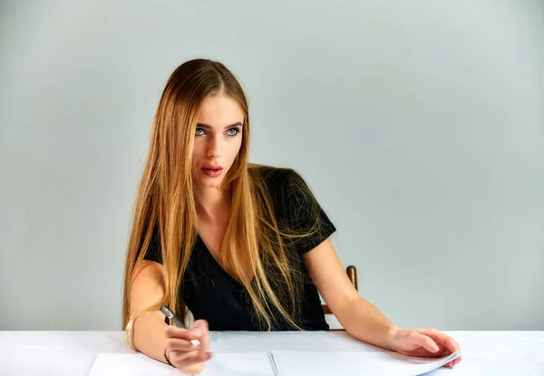 Retrato de una bonita chica rubia con el pelo largo y un gran maquillaje sobre un fondo blanco. Modelo estudiante manager sentado en diferentes poses en una mesa delante de la cámara en el estudio . — Foto de Stock