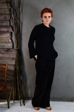 A woman is standing next to a chair right in front of the camera in various poses. Art portrait of a pretty model with short red hair in dark clothes on a gray background in the interior.