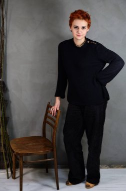 A woman is standing next to a chair right in front of the camera in various poses. Art portrait of a pretty model with short red hair in dark clothes on a gray background in the interior.