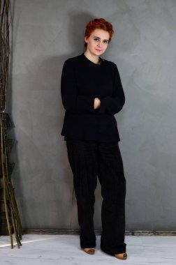 A woman is standing next to a chair right in front of the camera in various poses. Art portrait of a pretty model with short red hair in dark clothes on a gray background in the interior.