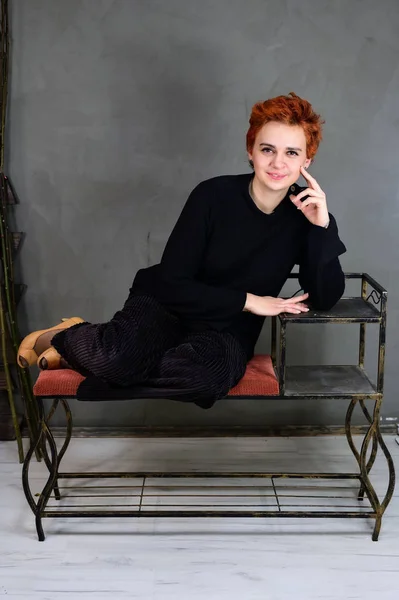 Kunstportret van een mooi model met kort rood haar in donkere kleren op een grijze achtergrond in het interieur. Een vrouw zit op een stoel vlak voor de camera in verschillende poses. — Stockfoto