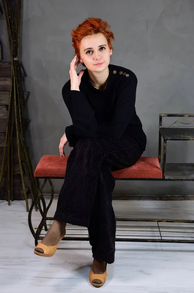 Kunstportret van een mooi model met kort rood haar in donkere kleren op een grijze achtergrond in het interieur. Een vrouw zit op een stoel vlak voor de camera in verschillende poses. — Stockfoto