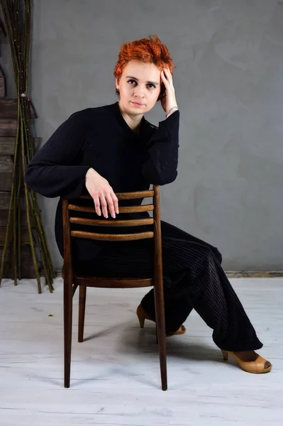 Kunstportret van een mooi model met kort rood haar in donkere kleren op een grijze achtergrond in het interieur. Een vrouw zit op een stoel vlak voor de camera in verschillende poses. — Stockfoto