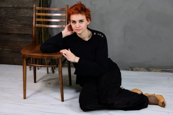 Portrait d'art d'un joli modèle aux cheveux courts roux en vêtements sombres sur fond gris à l'intérieur. Une femme assise à côté d'une chaise juste en face de la caméra dans différentes poses . — Photo