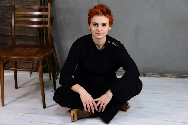 Portrait d'art d'un joli modèle aux cheveux courts roux en vêtements sombres sur fond gris à l'intérieur. Une femme assise à côté d'une chaise juste en face de la caméra dans différentes poses . — Photo