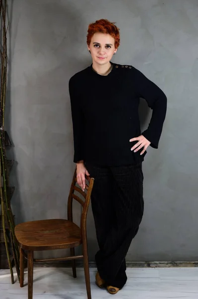 A woman is standing next to a chair right in front of the camera in various poses. Art portrait of a pretty model with short red hair in dark clothes on a gray background in the interior. — ストック写真