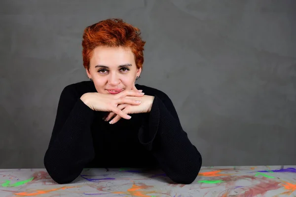 Une femme s'assoit à une table juste en face de la caméra dans diverses poses. Portrait d'art d'un joli modèle aux cheveux courts roux en vêtements sombres sur fond gris alternatif . — Photo