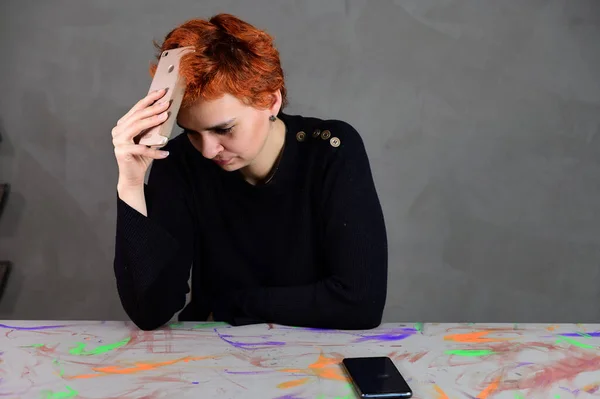 Μια γυναίκα χρησιμοποιεί ένα smartphone, κάθεται σε ένα τραπέζι ακριβώς μπροστά από την κάμερα σε διάφορες στάσεις. Πορτραίτο τέχνης ενός όμορφου μοντέλου με κοντά κόκκινα μαλλιά σε σκούρα ρούχα σε γκρι εναλλακτικό φόντο. — Φωτογραφία Αρχείου