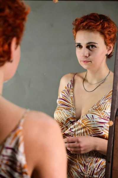 A woman poses in front of a mirror in front of the camera in various poses. Art portrait of a pretty model with short red hair in light overalls on a gray alternative background. — Stock Photo, Image