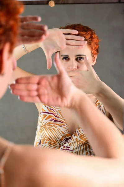 Μια γυναίκα ποζάρει μπροστά από έναν καθρέφτη μπροστά από την κάμερα σε διάφορες στάσεις. Πορτραίτο τέχνης ενός όμορφου μοντέλου με κοντά κόκκινα μαλλιά στο φως φόρμες σε ένα γκρι εναλλακτικό φόντο. — Φωτογραφία Αρχείου