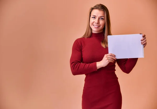 Retrato de una hermosa chica rubia con una hermosa sonrisa y excelentes dientes en un vestido de color burdeos con una carpeta en sus manos sobre un fondo rosa. Lindas miradas a la cámara, de pie recta . — Foto de Stock