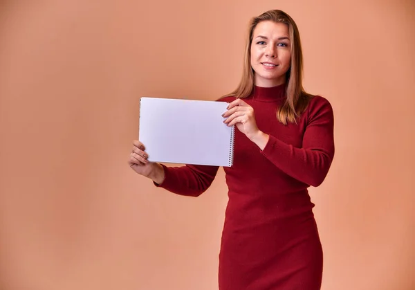 Retrato de una hermosa chica rubia con una hermosa sonrisa y excelentes dientes en un vestido de color burdeos con una carpeta en sus manos sobre un fondo rosa. Lindas miradas a la cámara, de pie recta . — Foto de Stock