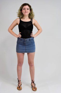 Siyah tişörtlü ve mavi etekli genç bir kadının yatay fotoğraf konsepti beyaz arka planda kameranın önünde duruyor. Güzel kıvırcık saçlı güzel bir kızın tam boy portresi..