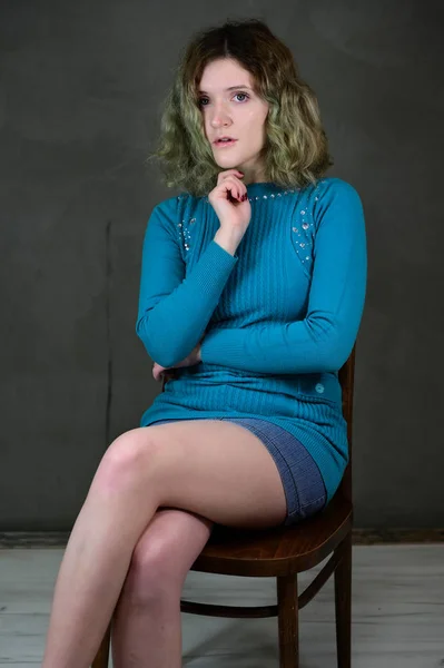 椅子に座って青いドレスで若い女性の概念垂直写真。ポートレートの美しいです学生の女の子とともに美しいです巻き毛カメラの前で灰色の背景. — ストック写真
