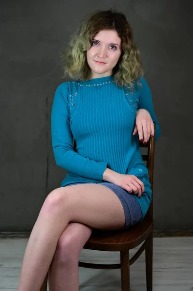 Onderwerp verticale foto van een jonge vrouw in een blauwe jurk zittend op een stoel. Portret van een mooi studentje met mooi krullend haar voor de camera op een grijze achtergrond. — Stockfoto