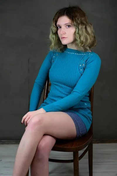 Концепція вертикального фото молодої жінки в блакитній сукні, що сидить на стільці. Портрет красивої студентки з красивим кучерявим волоссям перед камерою на сірому фоні . — стокове фото