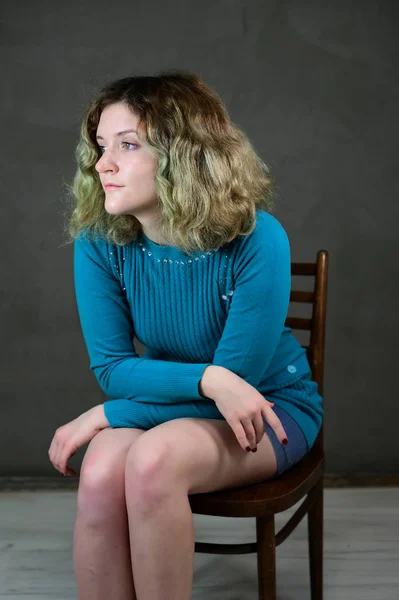 Begreppet vertikalt foto av en ung kvinna i en blå klänning sitter på en stol. Porträtt av en söt student flicka med vackra lockigt hår framför kameran på en grå bakgrund. — Stockfoto