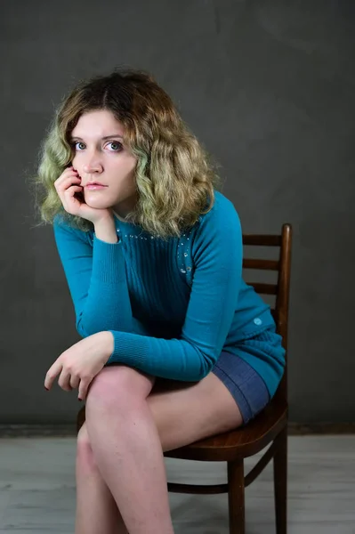 Begreppet vertikalt foto av en ung kvinna i en blå klänning sitter på en stol. Porträtt av en söt student flicka med vackra lockigt hår framför kameran på en grå bakgrund. — Stockfoto