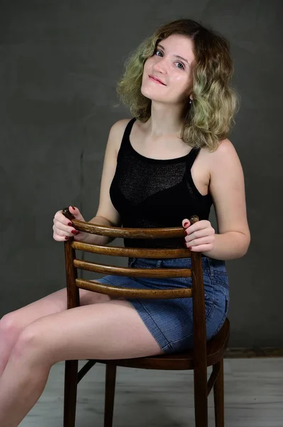 Retrato de una bonita estudiante con hermoso pelo rizado delante de la cámara sobre un fondo gris. Concepto foto vertical de una mujer joven con una camiseta negra y falda azul sentada en una silla . — Foto de Stock