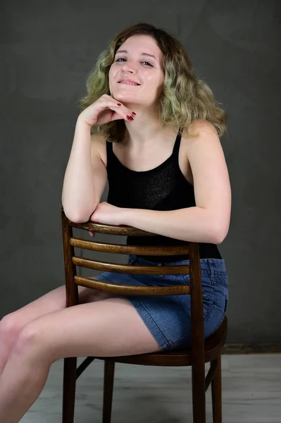 Portret van een mooi studentmeisje met mooi krullend haar voor de camera op een grijze achtergrond. Onderwerp verticale foto van een jonge vrouw in een zwart T-shirt en blauwe rok zittend op een stoel. — Stockfoto