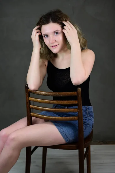 Portrait of a pretty student girl with beautiful curly hair in front of the camera on a gray background. Concept vertical photo of a young woman in a black T-shirt and blue skirt sitting on a chair. — 스톡 사진