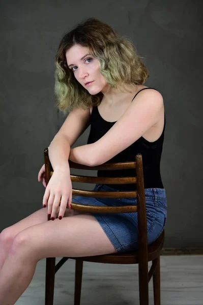 Retrato de una bonita estudiante con hermoso pelo rizado delante de la cámara sobre un fondo gris. Concepto foto vertical de una mujer joven con una camiseta negra y falda azul sentada en una silla . — Foto de Stock