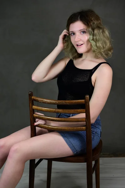 Porträt eines hübschen Studentenmädchens mit schönen lockigen Haaren vor der Kamera auf grauem Hintergrund. Konzeptfoto einer jungen Frau in schwarzem T-Shirt und blauem Rock, die auf einem Stuhl sitzt. — Stockfoto