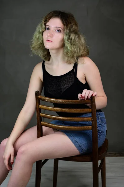 Porträtt av en söt student flicka med vackra lockigt hår framför kameran på en grå bakgrund. Begreppet vertikalt foto av en ung kvinna i svart T-shirt och blå kjol sitter på en stol. — Stockfoto