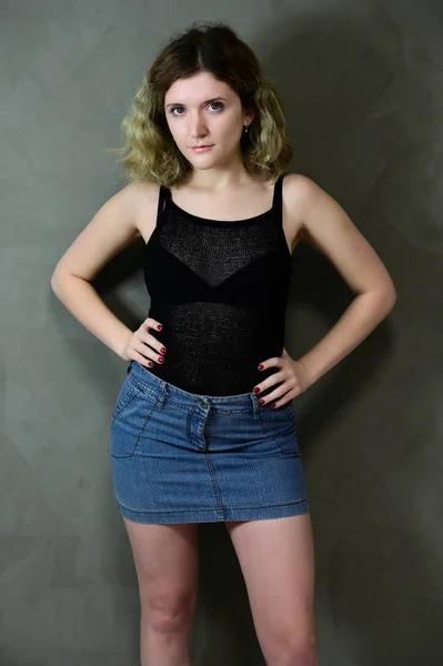 Concepto de foto vertical de una mujer joven con una camiseta negra y falda azul de pie recta. Retrato de una bonita estudiante con hermoso pelo rizado delante de la cámara sobre un fondo gris . —  Fotos de Stock