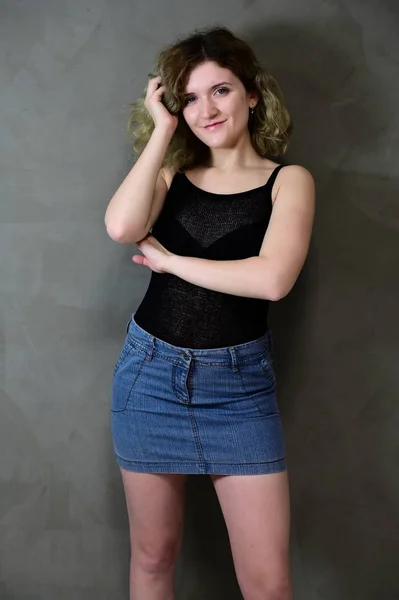 Concept vertical photo of a young woman in a black T-shirt and blue skirt standing straight. Portrait of a pretty student girl with beautiful curly hair in front of the camera on a gray background. — 스톡 사진