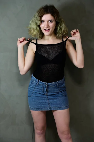 Concept vertical photo of a young woman in a black T-shirt and blue skirt standing straight. Portrait of a pretty student girl with beautiful curly hair in front of the camera on a gray background. — Stock Photo, Image