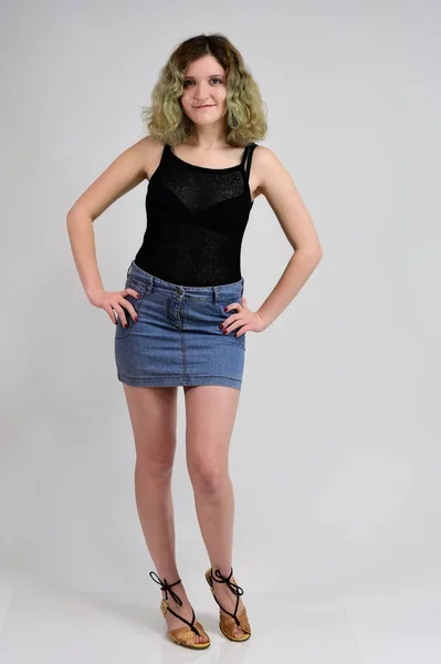 Un concept photo horizontal d'une jeune femme en t-shirt noir et jupe bleue est debout devant l'appareil photo sur un fond blanc. Portrait complet d'une jolie fille aux beaux cheveux bouclés . — Photo