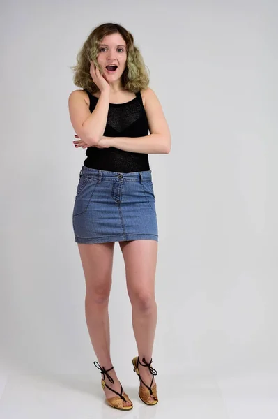 Ein horizontales Fotokonzept einer jungen Frau in schwarzem T-Shirt und blauem Rock steht vor der Kamera auf weißem Hintergrund. Ganzkörperporträt eines hübschen Mädchens mit schönen lockigen Haaren. — Stockfoto