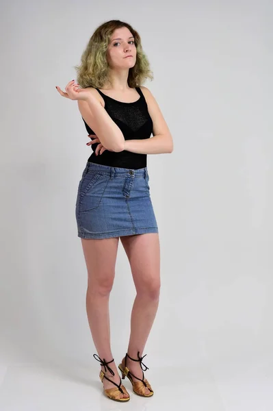 A horizontal photo concept of a young woman in a black t-shirt and blue skirt is standing in front of the camera on a white background. Full-length portrait of a pretty girl with beautiful curly hair. — 스톡 사진