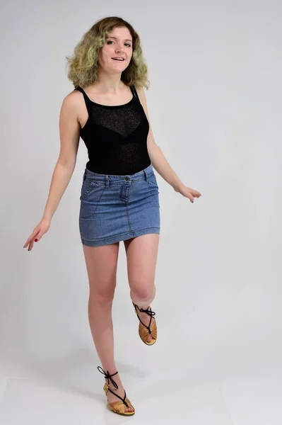 A horizontal photo concept of a young woman in a black t-shirt and blue skirt is standing in front of the camera on a white background. Full-length portrait of a pretty girl with beautiful curly hair. — Stock Photo, Image