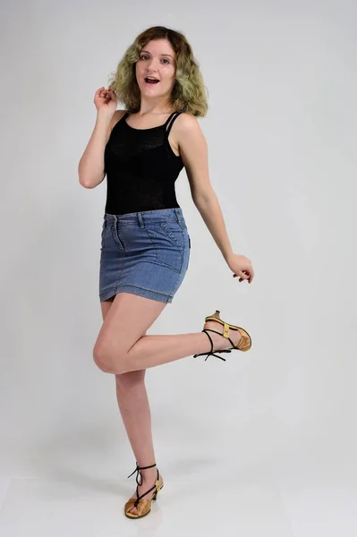 Um conceito de foto horizontal de uma jovem mulher em uma camiseta preta e saia azul está de pé na frente da câmera em um fundo branco. Retrato de comprimento total de uma menina bonita com cabelo encaracolado bonito . — Fotografia de Stock