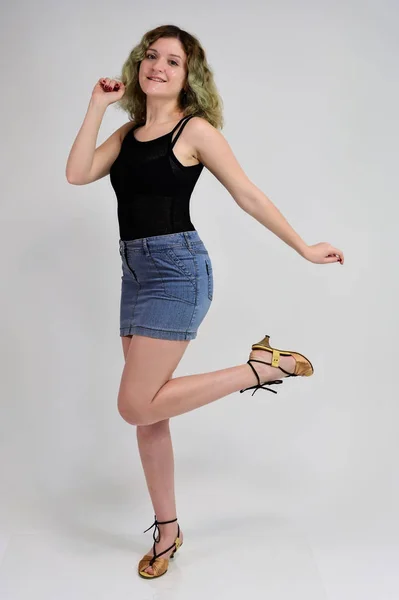 A horizontal photo concept of a young woman in a black t-shirt and blue skirt is standing in front of the camera on a white background. Full-length portrait of a pretty girl with beautiful curly hair. — Stock Photo, Image