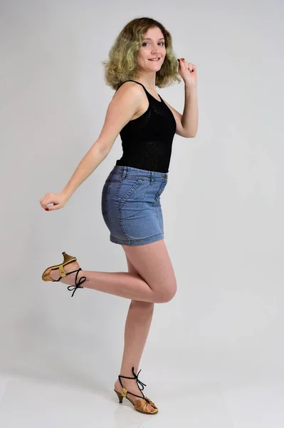 Um conceito de foto horizontal de uma jovem mulher em uma camiseta preta e saia azul está de pé na frente da câmera em um fundo branco. Retrato de comprimento total de uma menina bonita com cabelo encaracolado bonito . — Fotografia de Stock