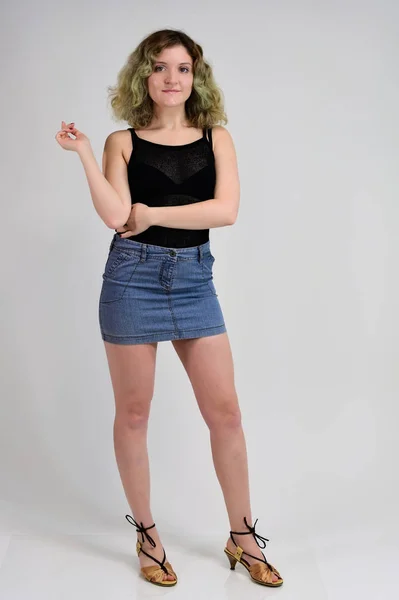 Ein horizontales Fotokonzept einer jungen Frau in schwarzem T-Shirt und blauem Rock steht vor der Kamera auf weißem Hintergrund. Ganzkörperporträt eines hübschen Mädchens mit schönen lockigen Haaren. — Stockfoto