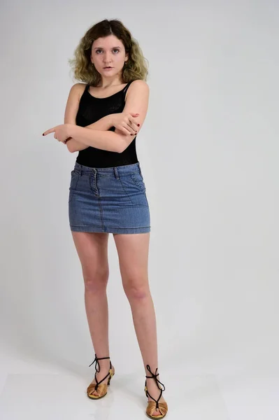 Full-length portrait of a pretty girl with beautiful curly hair. A horizontal photo concept of a young woman in a black t-shirt and blue skirt is standing in front of the camera on a white background. — Stock Photo, Image