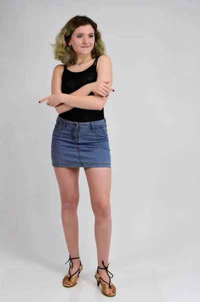 Full-length portrait of a pretty girl with beautiful curly hair. A horizontal photo concept of a young woman in a black t-shirt and blue skirt is standing in front of the camera on a white background. — 스톡 사진