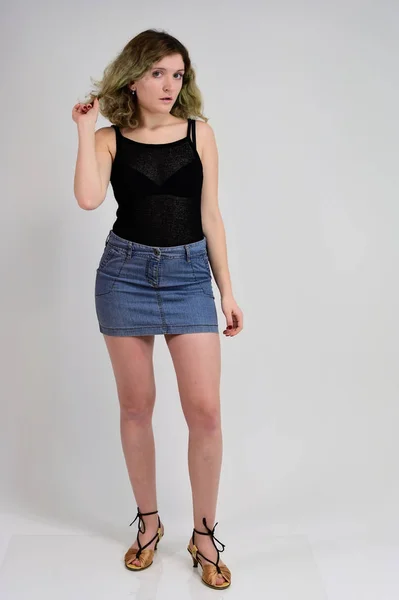 Full-length portrait of a pretty girl with beautiful curly hair. A horizontal photo concept of a young woman in a black t-shirt and blue skirt is standing in front of the camera on a white background. — 스톡 사진