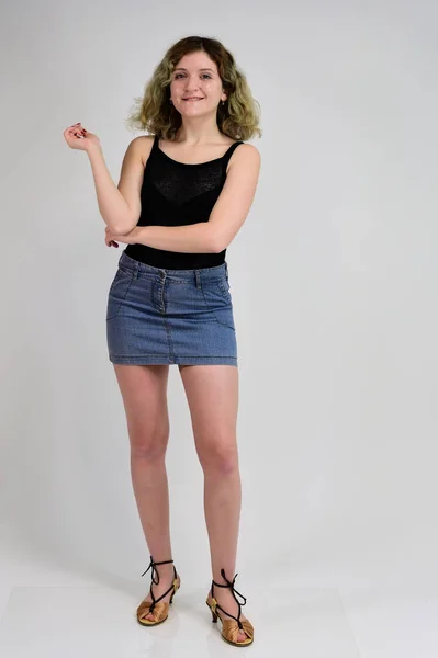 Retrato de larga duración de una chica bonita con hermoso cabello rizado. Un concepto de fotografía horizontal de una mujer joven con una camiseta negra y una falda azul está de pie frente a la cámara sobre un fondo blanco . — Foto de Stock