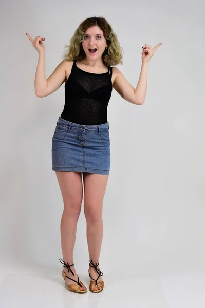 Full-length portrait of a pretty girl with beautiful curly hair. A horizontal photo concept of a young woman in a black t-shirt and blue skirt is standing in front of the camera on a white background. — 스톡 사진