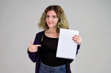 Siyah ceketli, beyaz arka planda kameranın önünde duran genç bir kadının yatay fotoğrafını hayal et. Elinde dosyasıyla pofuduk saçlı bir kız öğrencinin portresi..