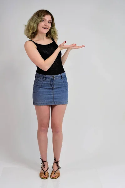 Full-length portrait of a pretty girl with beautiful curly hair. A horizontal photo concept of a young woman in a black t-shirt and blue skirt is standing in front of the camera on a white background. — 스톡 사진