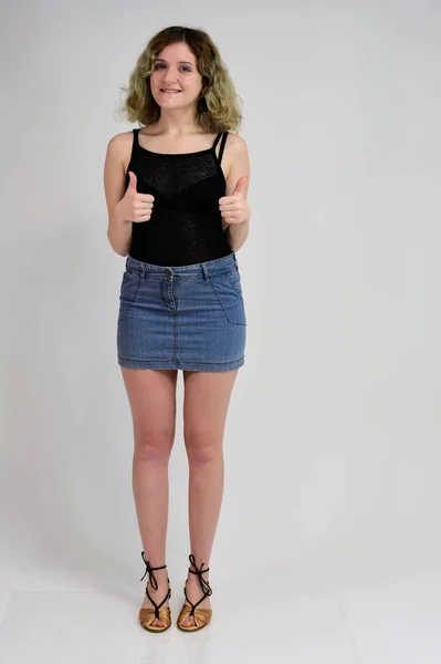 Full-length portrait of a pretty girl with beautiful curly hair. A horizontal photo concept of a young woman in a black t-shirt and blue skirt is standing in front of the camera on a white background. — 스톡 사진