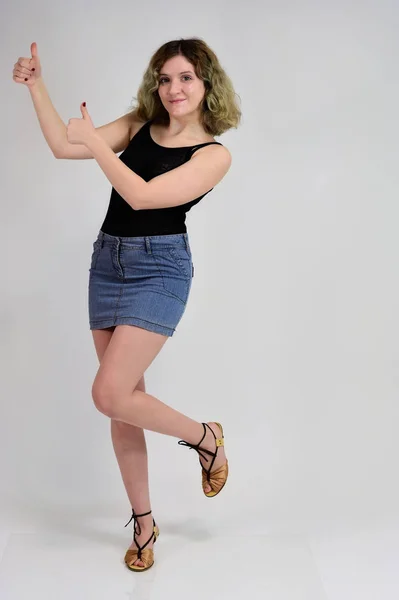 Full-length portrait of a pretty girl with beautiful curly hair. A horizontal photo concept of a young woman in a black t-shirt and blue skirt is standing in front of the camera on a white background. — 스톡 사진