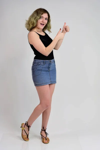 Full-length portrait of a pretty girl with beautiful curly hair. A horizontal photo concept of a young woman in a black t-shirt and blue skirt is standing in front of the camera on a white background. — 스톡 사진