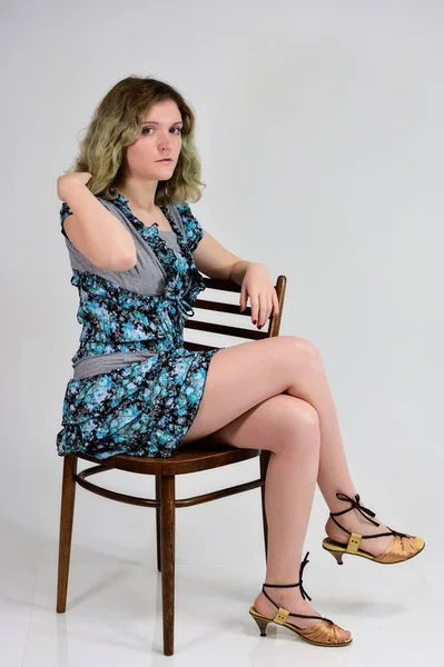 Begreppet vertikalt foto av en ung kvinna i en turkos klänning sitter på en stol framför kameran på en vit bakgrund. Porträtt av en student flicka med vackra magnifika hår. — Stockfoto